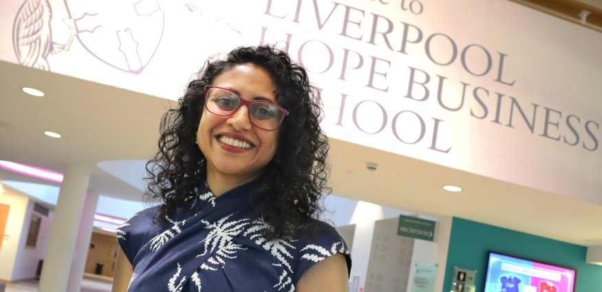 A woman wearing glasses and a blue and white dress stands in front of a wall with a Liverpool Hope Business School sign.